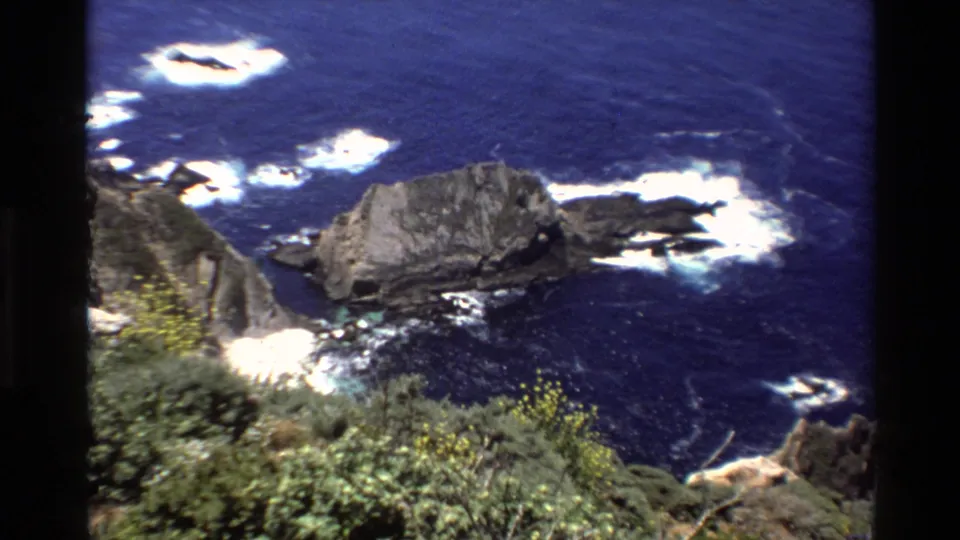 1980 home-movie frame of the Big Sur shoreline, with a large rock formation, white surf patterns, dark blue water, and blurred cliffside plants.