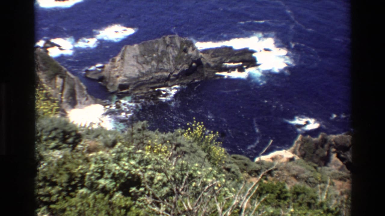 1980 8mm Big Sur view looking down over coastal shrubs, rock formations, and waves breaking in deep blue water.