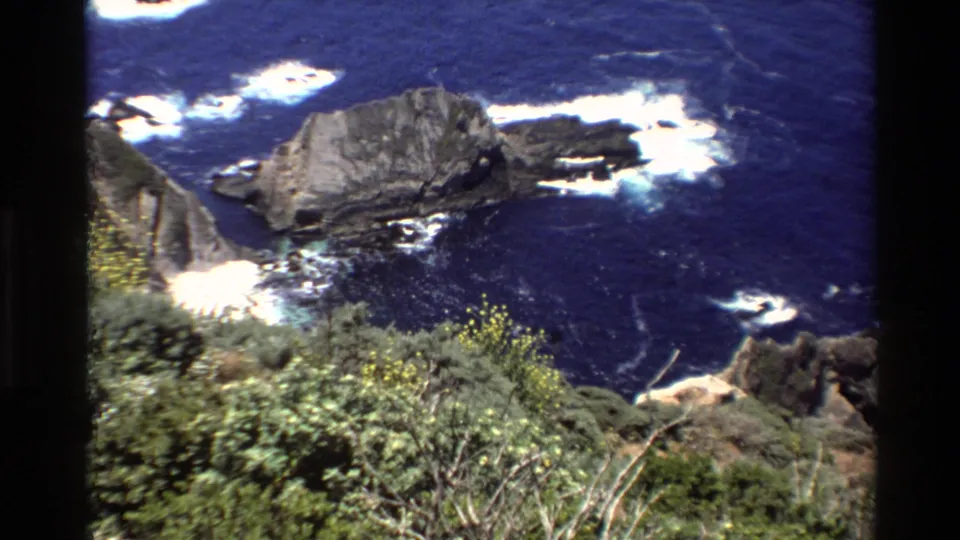 1980 8mm Big Sur view looking down over coastal shrubs, rock formations, and waves breaking in deep blue water.