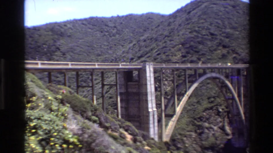 1980 Big Sur travel footage frame showing a concrete arch bridge spanning a green, steep-sided canyon.