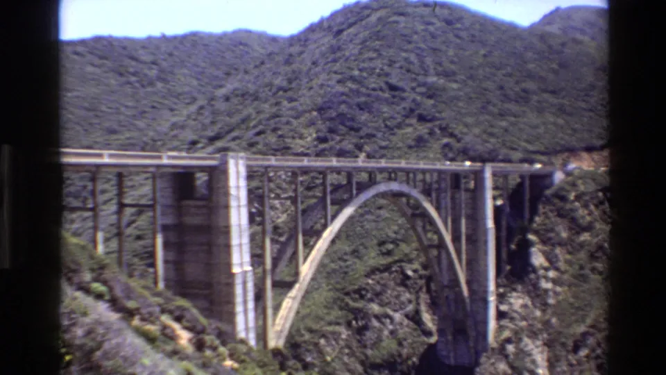 Big Sur 8mm frame of a concrete arch bridge across a mountain canyon, with hillsides and rock visible around it.