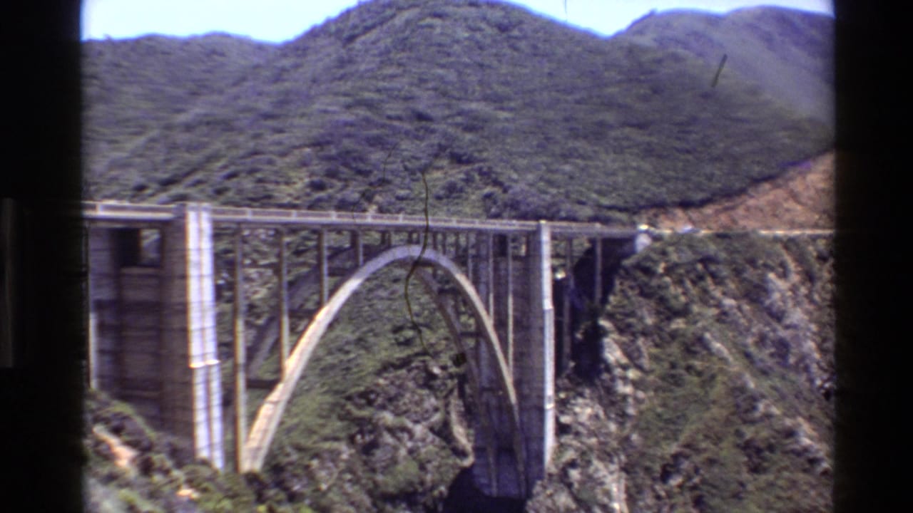 1980 Big Sur home-movie frame showing a large concrete arch bridge crossing a steep canyon below green coastal hills.