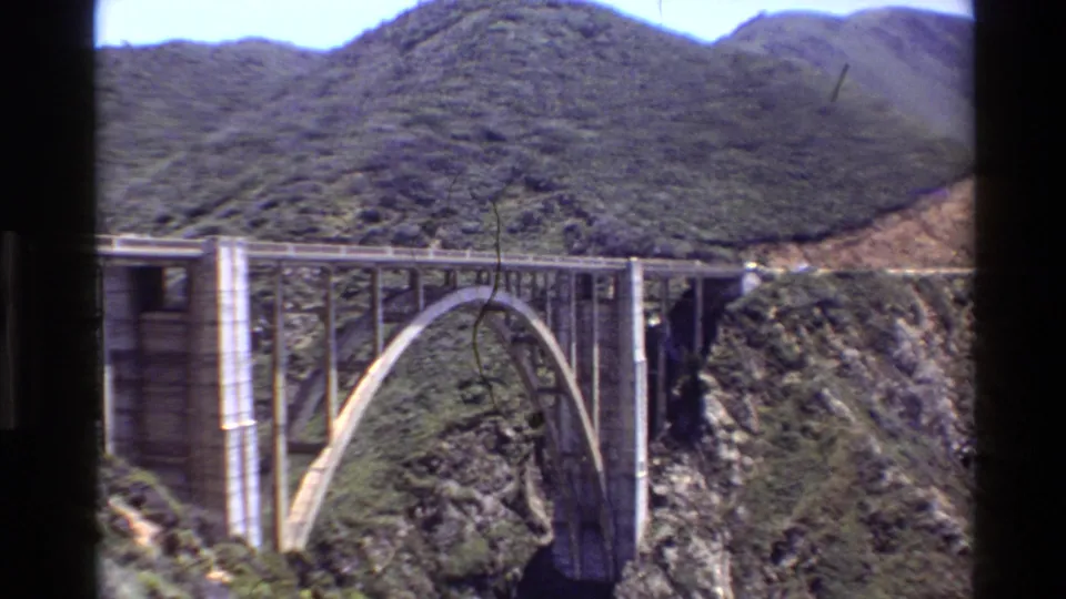 1980 Big Sur home-movie frame showing a large concrete arch bridge crossing a steep canyon below green coastal hills.