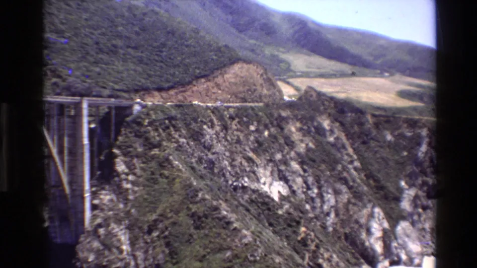 1980 Big Sur travel footage frame with the bridge edge at left, rugged canyon rock, hillside vegetation, and open terrain beyond.