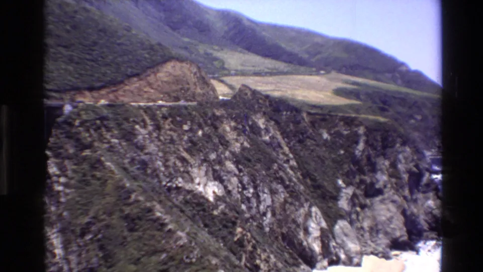 Big Sur 1980 home-movie frame showing rugged canyon terrain, green hillsides, and a sliver of bridge structure at the edge.