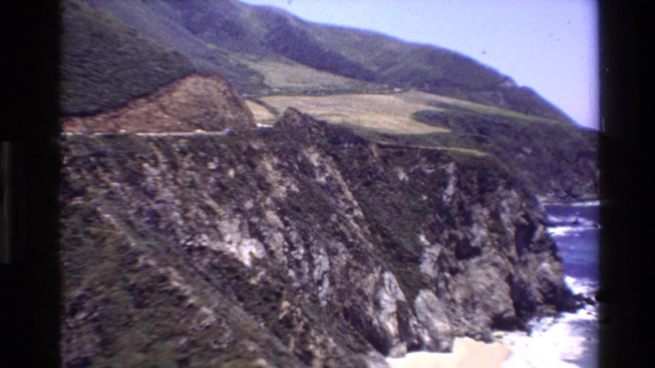 1980 8mm view of Big Sur cliffs dropping toward a small sandy beach with ocean surf below.