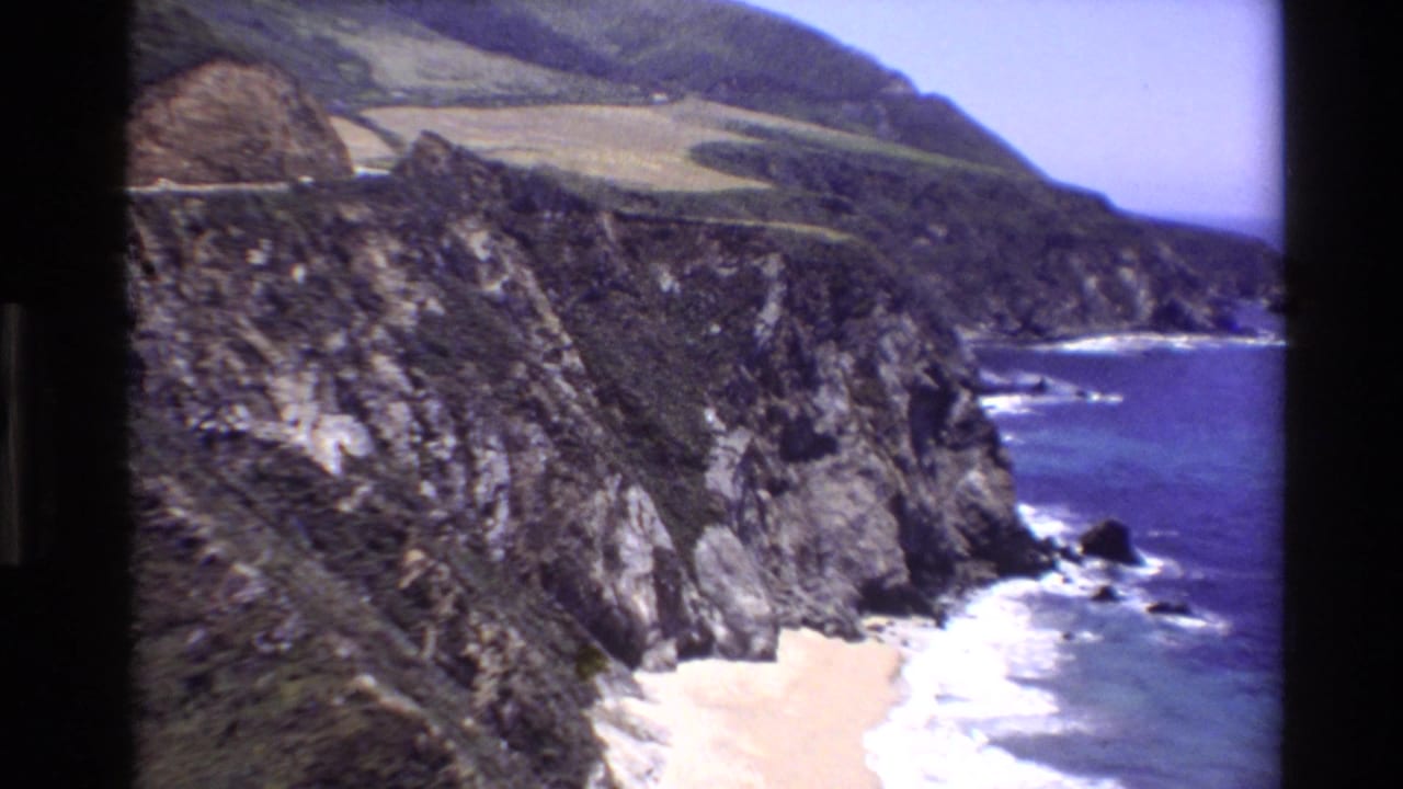 Coastal cliffs, hillside fields, ocean, and surf along a rocky shore.