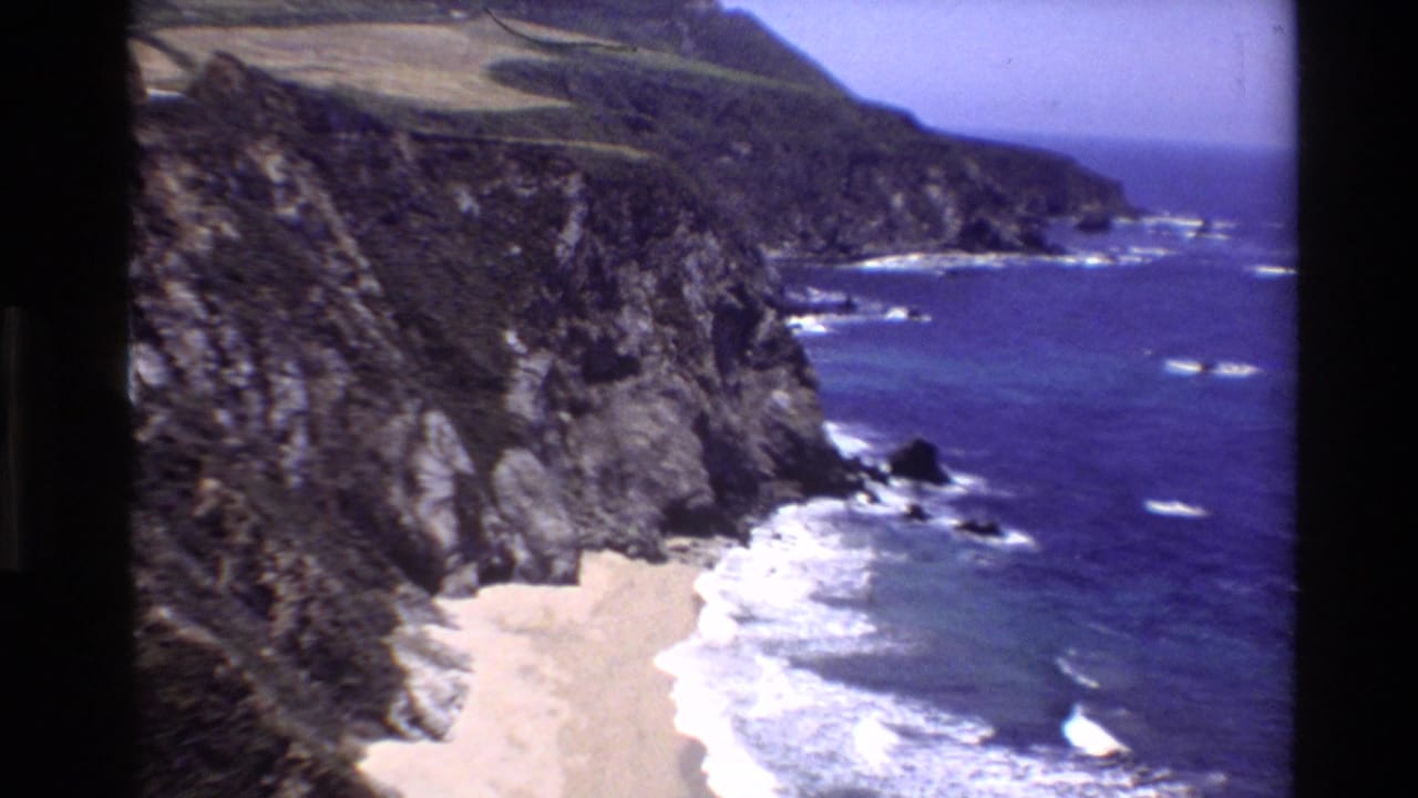 1980 Big Sur 8mm frame showing a sandy cove beneath rugged cliffs and deep blue Pacific water.