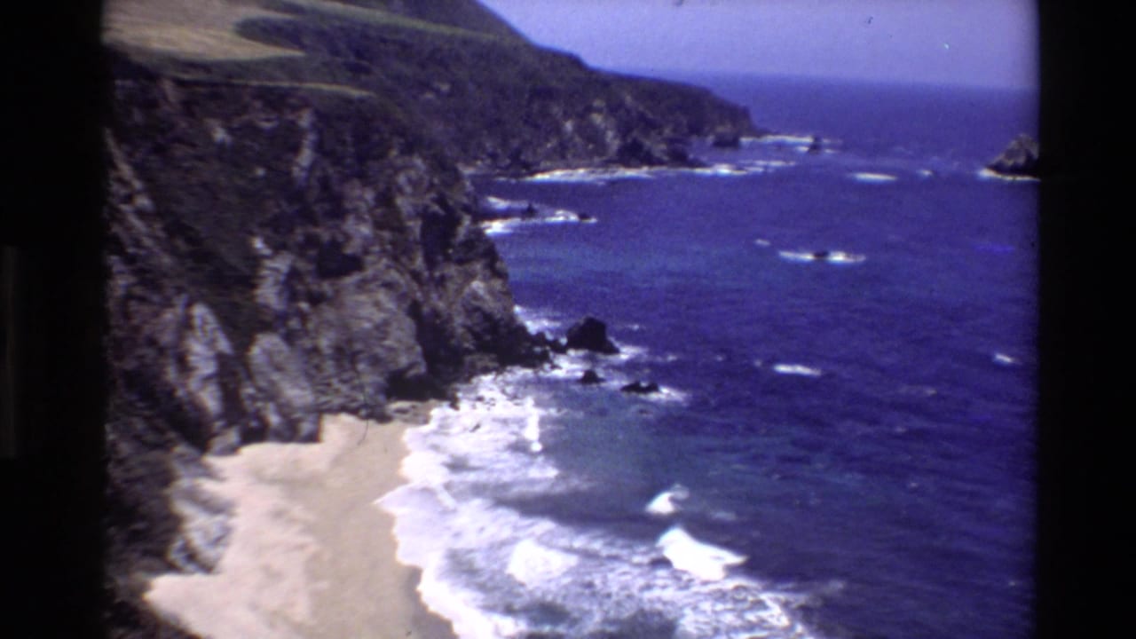 Big Sur coastal panorama from 1980 8mm film, showing cliffs, beach, offshore rocks, and surf.