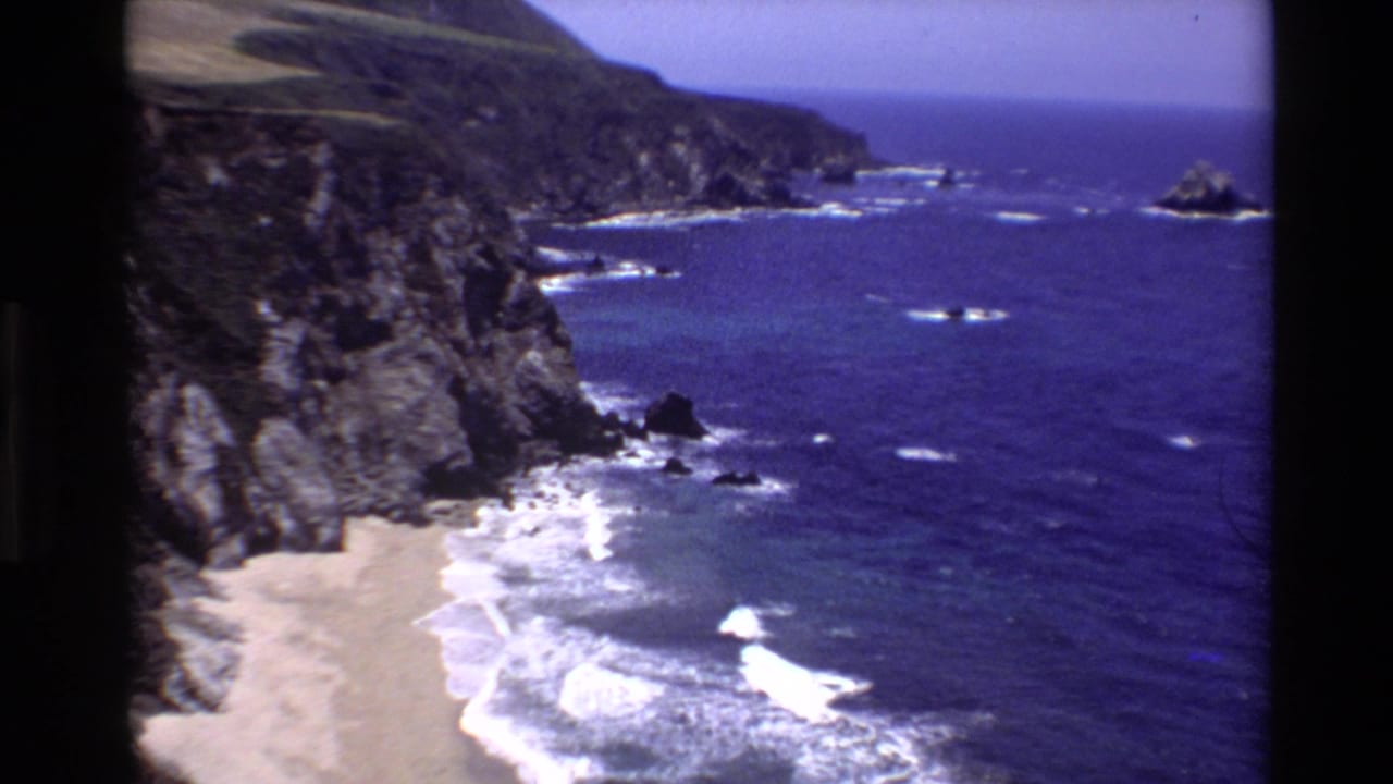 High coastal view of cliffs, sandy beach, white surf, and blue ocean.