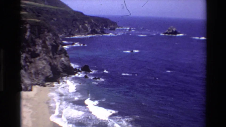 1980 8mm frame of the Big Sur coast showing dark cliffs, blue Pacific water, white wave breaks, and rocky outcrops offshore.