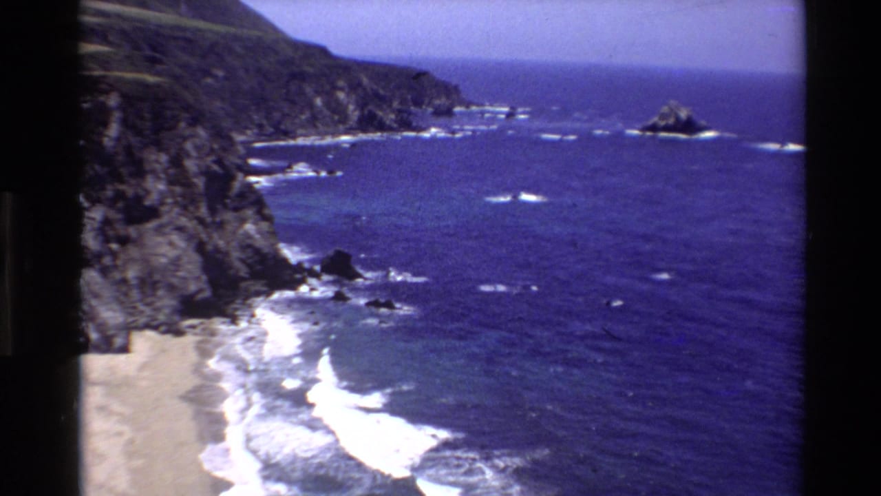 Big Sur home-movie frame from 1980 with cliff faces, a pale beach edge, white surf, and a rocky point extending into the Pacific.