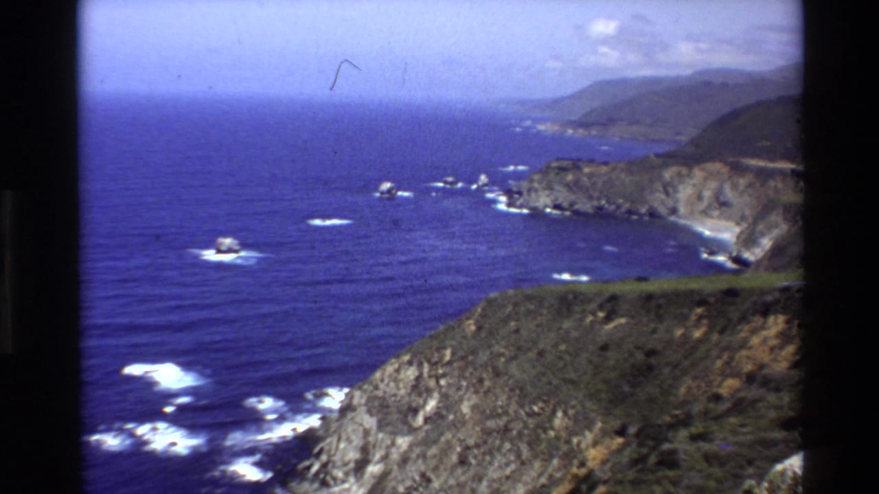 1980 8mm wide view of Big Sur headlands and Pacific water, with rocky foreground slopes, surf patches, and distant coastal hills.