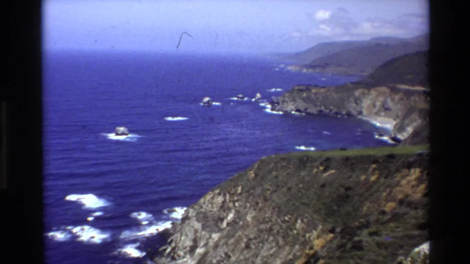 1980 8mm wide view of Big Sur headlands and Pacific water, with rocky foreground slopes, surf patches, and distant coastal hills.
