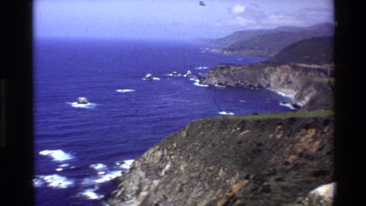 Rocky Big Sur coastline with blue ocean, white surf, cliffs, and distant hills.