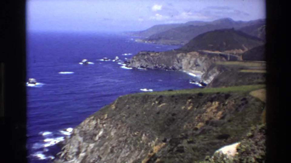 1980 home-movie frame of Big Sur cliffs, a winding coastal road, blue ocean, white surf, and layered hills in the distance.