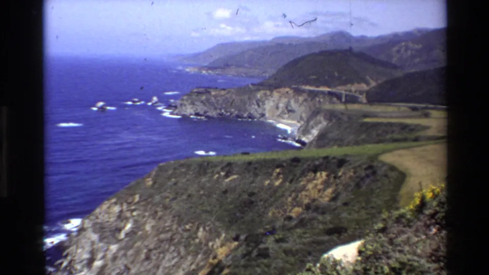 Wide 1980 8mm view over Big Sur’s rocky shore, Pacific surf, green bluff, coastal road, and hazy hills.