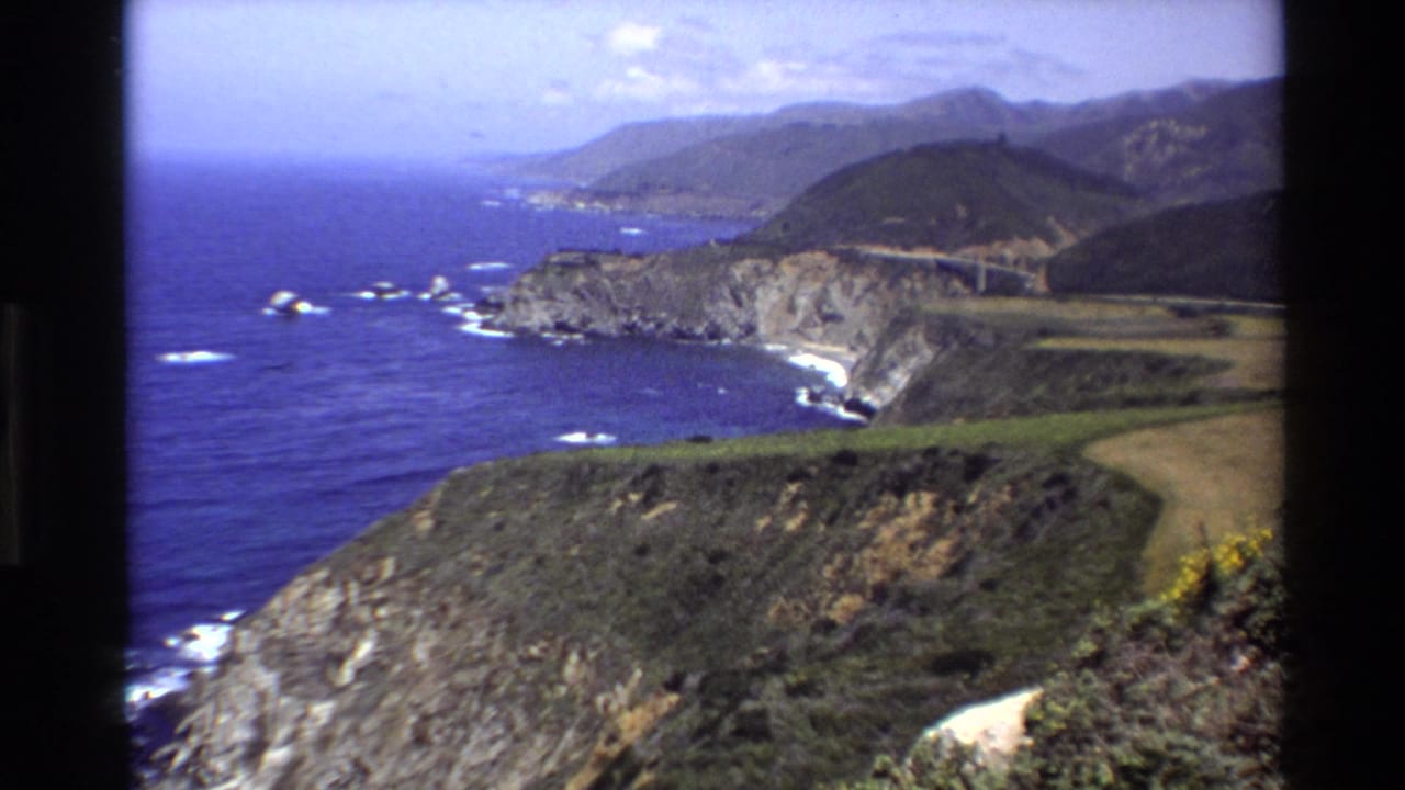 High coastal view of Big Sur cliffs, blue ocean, offshore rocks, and a winding road.