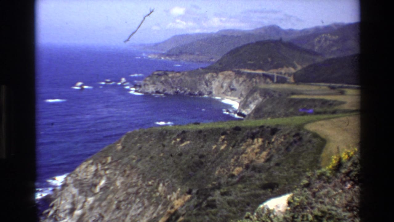 Coastal panorama with cliffs, ocean, hills, road, surf, and visible film dust.