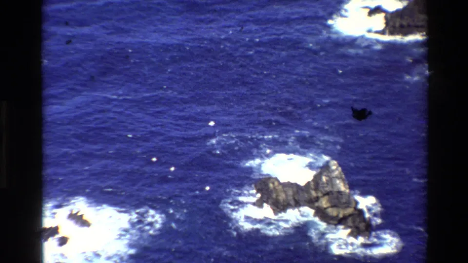 1980 8mm Big Sur ocean view focused downward on a rocky sea stack surrounded by white Pacific surf.