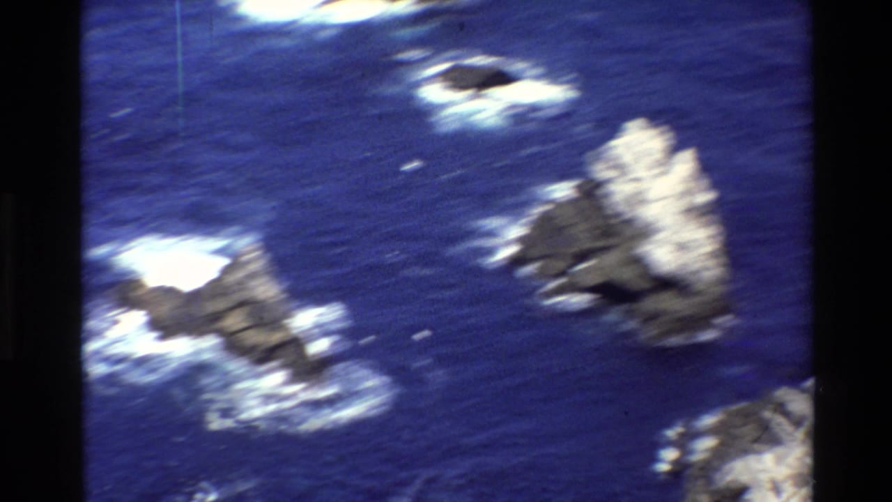 1980 Big Sur 8mm aerial view of offshore rocks in blue water, with motion blur and white surf around the sea stacks.