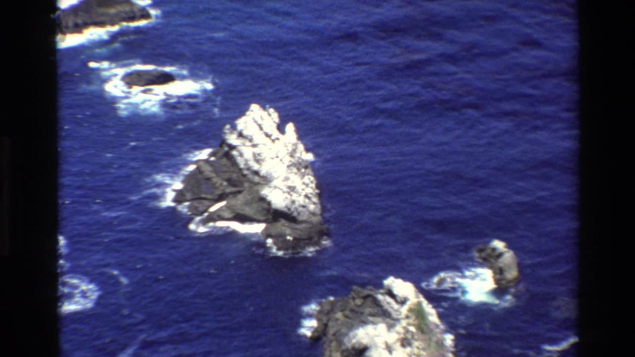 Jagged sea stack surrounded by blue ocean and white breaking waves.