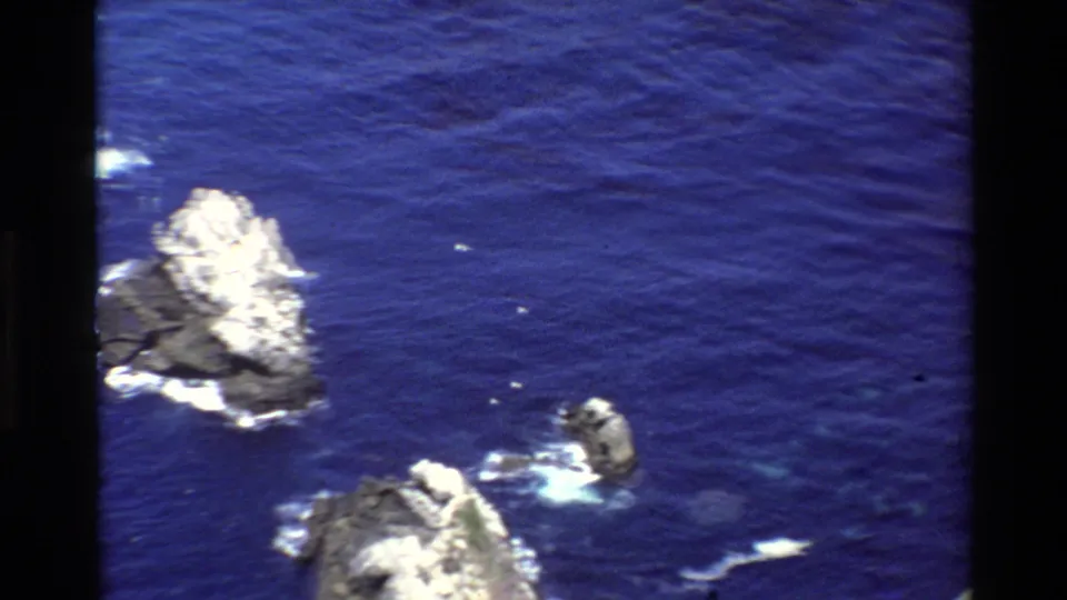 1980 Big Sur 8mm aerial view of multiple offshore rocks, white wave wash, and saturated blue ocean from a moving camera.