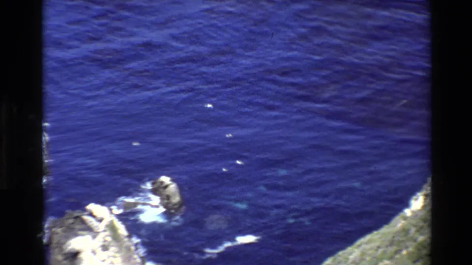 1980 Big Sur aerial frame with open blue water, small offshore rocks, white surf traces, and coastline visible at the lower right edge.