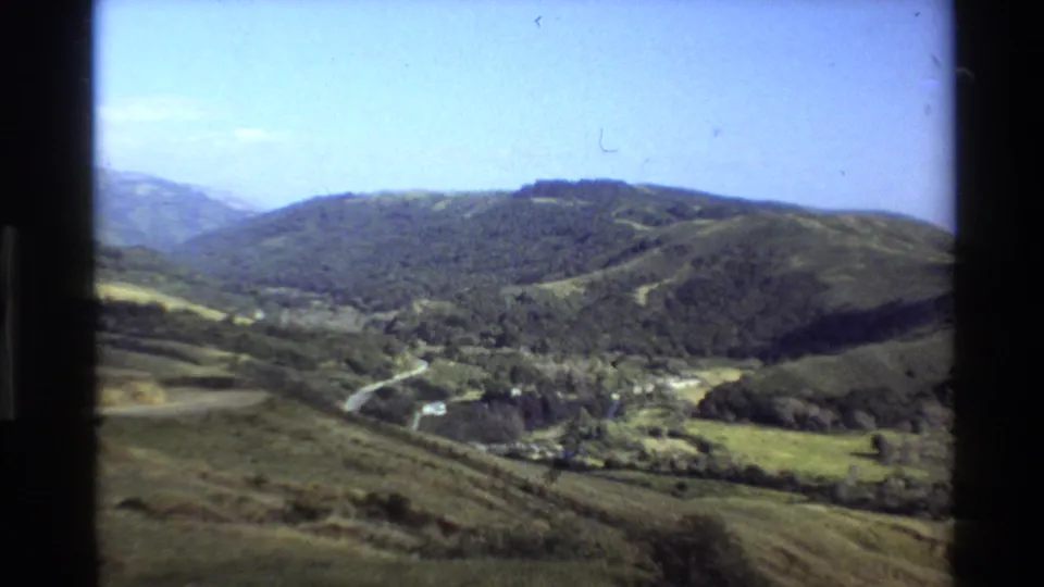1980 Big Sur landscape view with green hills, wooded slopes, a pale winding road, and clear blue sky.