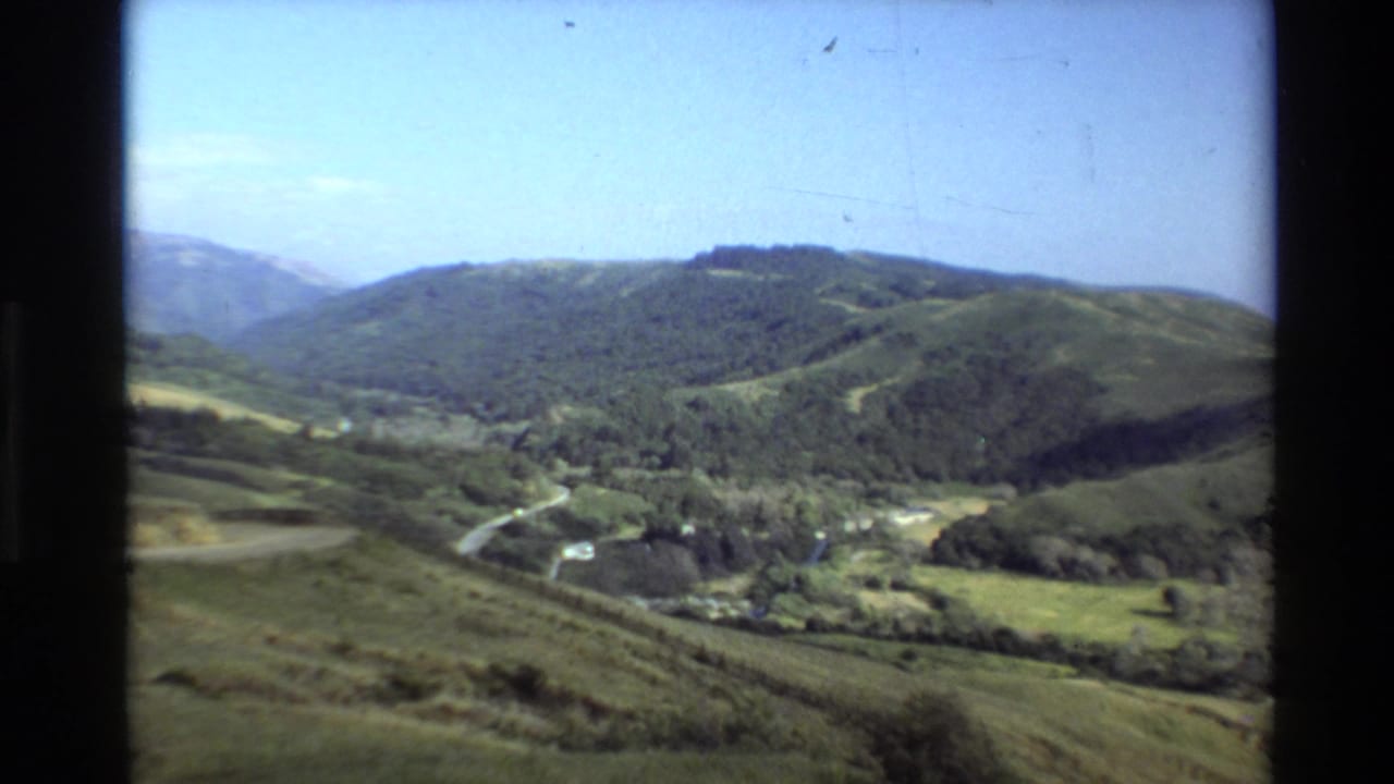 Soft 8mm view of Big Sur green hills, distant ridges, valley vegetation, and a road crossing the landscape.