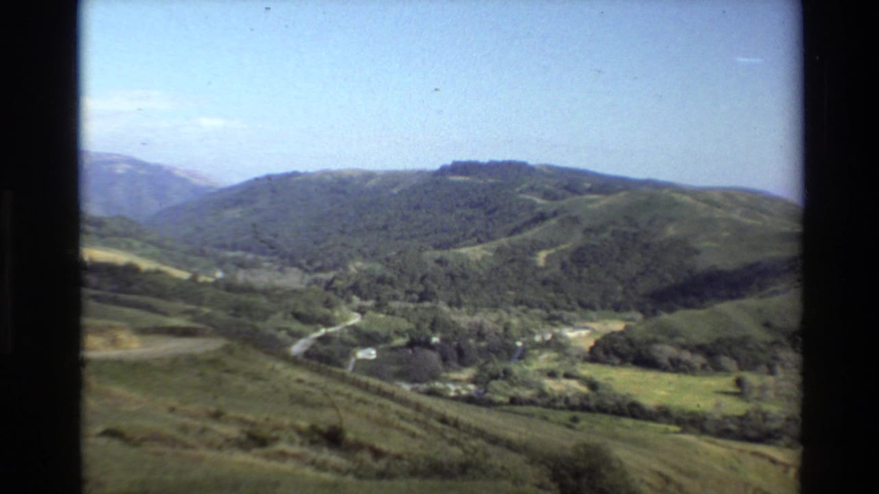Wide 1980 Big Sur home-movie frame showing rolling green hills, a pale road, trees, and bright blue sky.