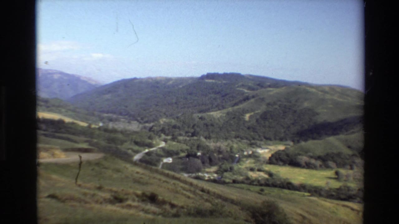 1980 8mm Big Sur view of green hills, a winding valley road, distant mountains, and a pale blue sky.