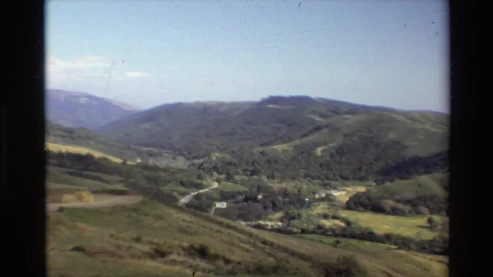 Big Sur, California 8mm frame showing forested hills, grassy slopes, a valley road, and distant mountain ridges.