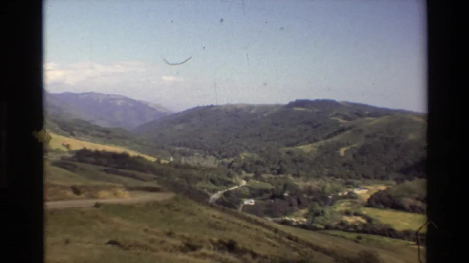 Vintage 8mm view of Big Sur hills and a valley road, with soft color, dust specks, and distant mountains.