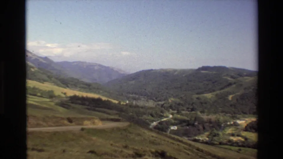 1980 Big Sur home-movie frame looking across green hills, wooded valley land, a winding road, and hazy mountain ridges.