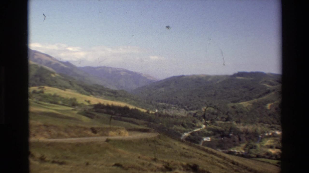 1980 Big Sur home-movie frame showing rolling hills, a forested valley, distant mountains, and a winding road under blue sky.