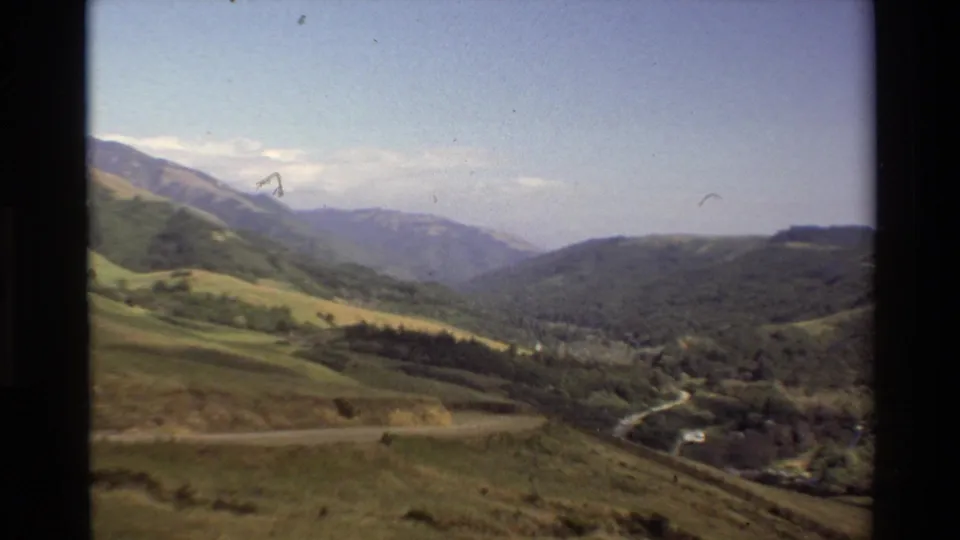 Big Sur valley view with green slopes, evergreen cover, a winding road, distant ridges, and partly cloudy blue sky.