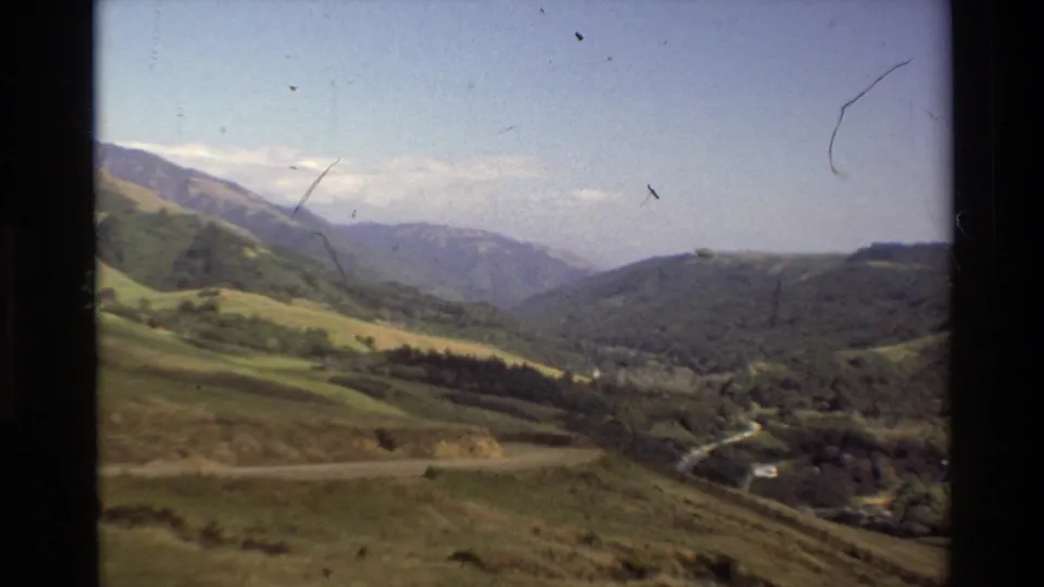 1980 Big Sur landscape frame showing layered hills, a forested valley, a visible road, and distant mountain ridges in soft 8mm focus.