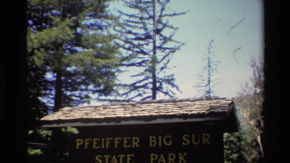 Bright 8mm frame showing the Pfeiffer Big Sur State Park sign with yellow lettering, roof shingles, trees, and sky.