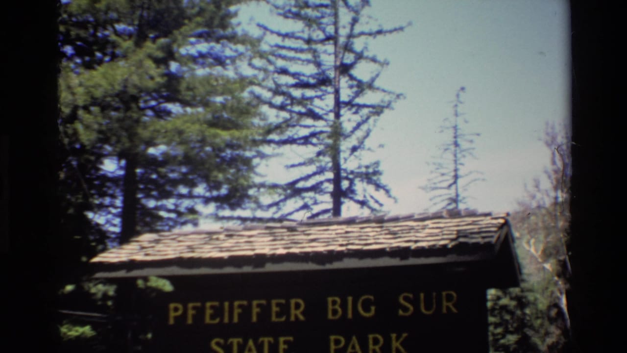 1980 8mm view of the Pfeiffer Big Sur State Park sign with a shingled roof, tall trees, and bright sky in Big Sur, California.
