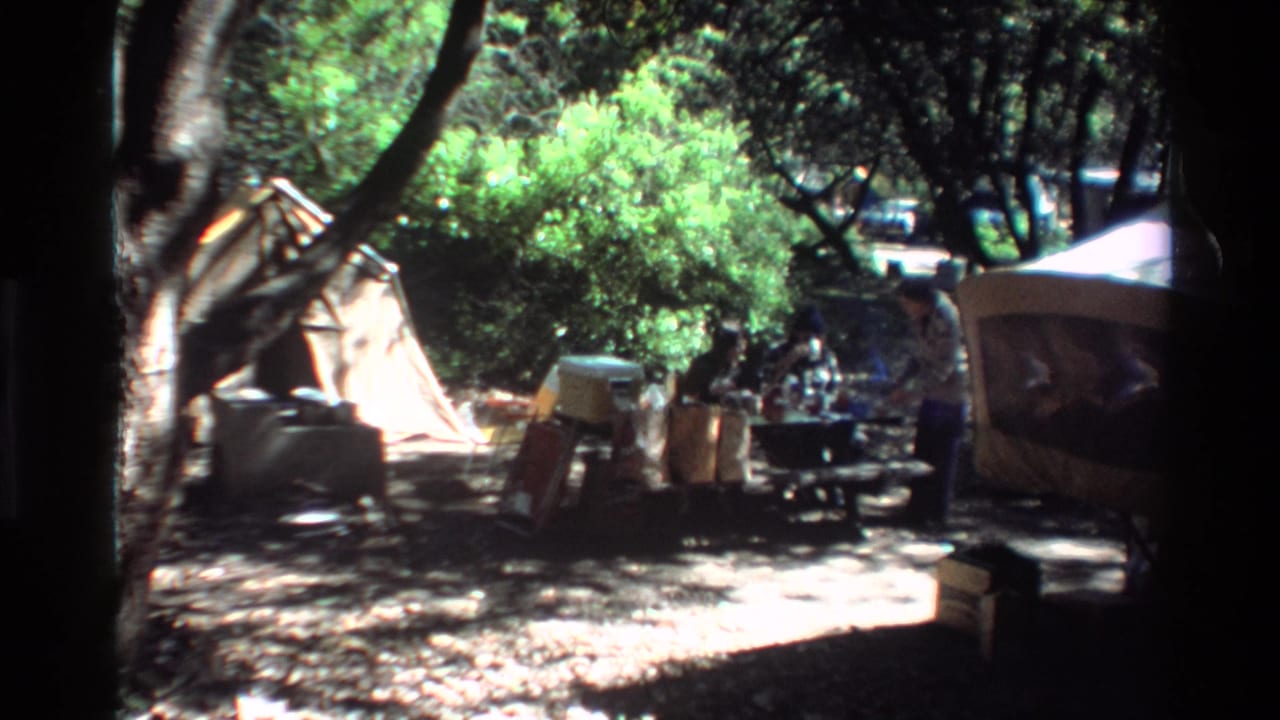 Campsite under trees with tent, picnic table, coolers, bags, and indistinct people nearby.