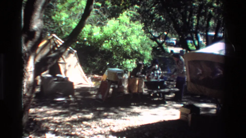 Wooded Big Sur camping scene from 1980 showing a tent, picnic table, coolers, supplies, and campers in shade.