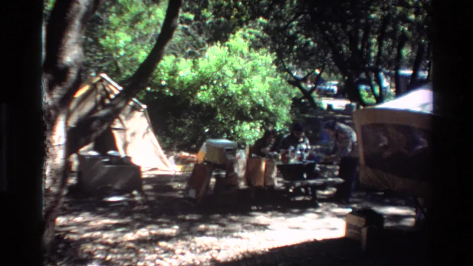 1980 Big Sur campsite view with a large tree trunk, canvas tent, chairs, coolers, and people partly hidden near a picnic table.