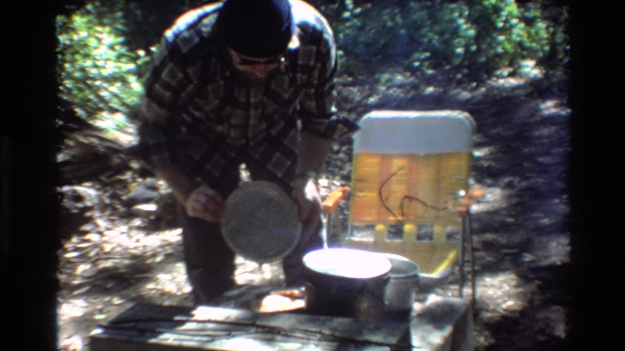Man in plaid shirt and dark beanie holds a pan near pots and a yellow chair outdoors.