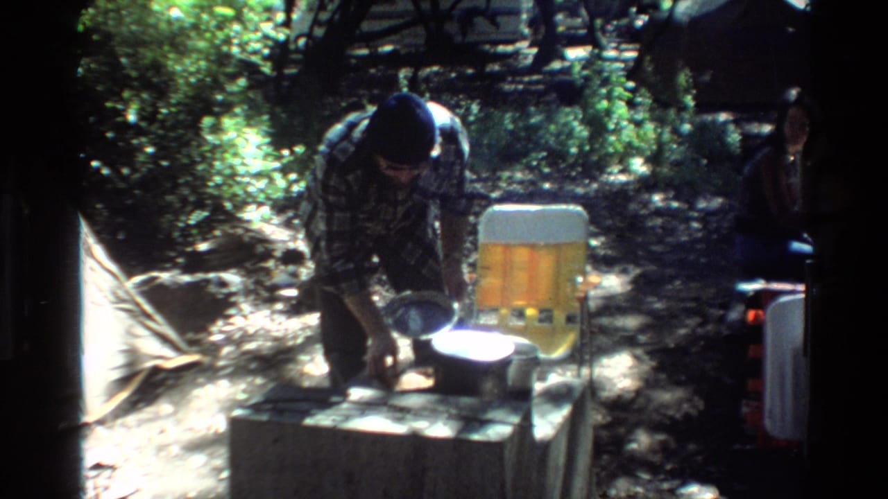 Big Sur campsite frame from 1980 showing a man leaning over pots on a table, with a yellow folding chair and another adult partly visible.