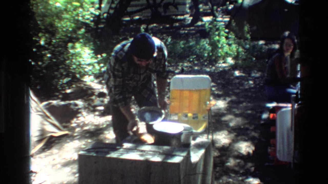 1980 Big Sur home-movie frame with a man working over campsite cookware beside a yellow chair and a seated adult in the background.