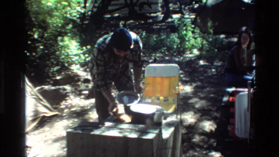1980 Big Sur home-movie frame with a man working over campsite cookware beside a yellow chair and a seated adult in the background.