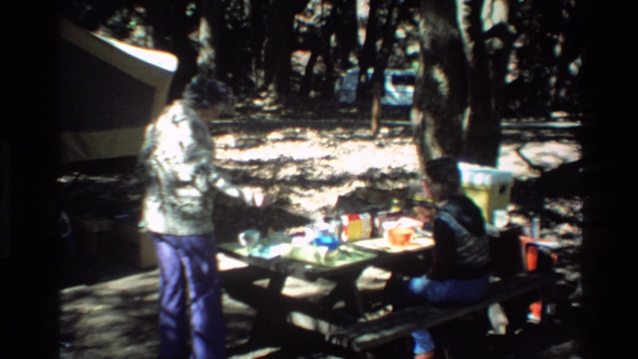 Two adults at a picnic table in a shaded wooded campsite.