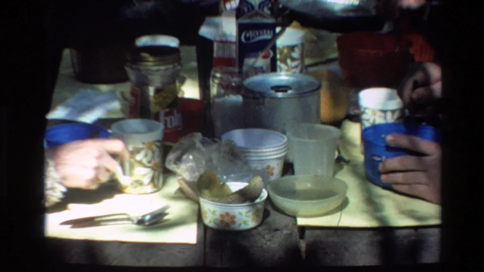 Big Sur 1980 campsite table detail with cups, utensils, floral bowls, containers, and adult hands handling food or drink.