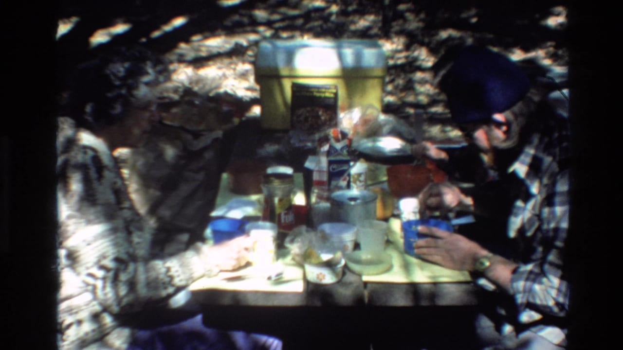 Big Sur 1980 home-movie frame showing two adults at a campsite table with dishes, cups, containers, and a coffee can.